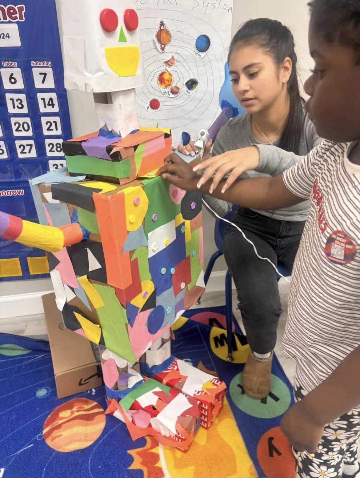 Preschoolers add pieces of colored paper to a robot made of cardboard during a STEM activity.