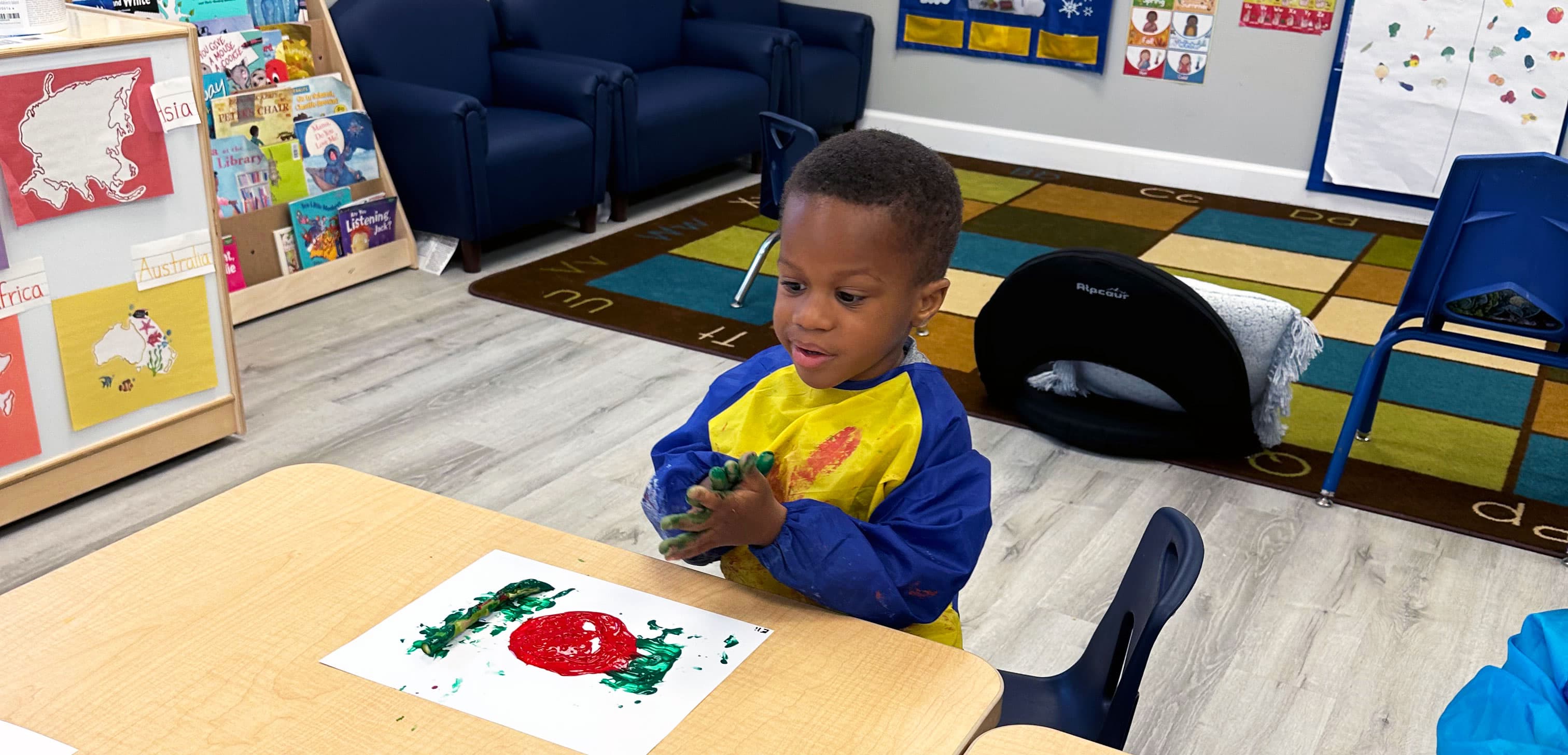 A child in a paint smock smiling at the apple print they made with green and red paint