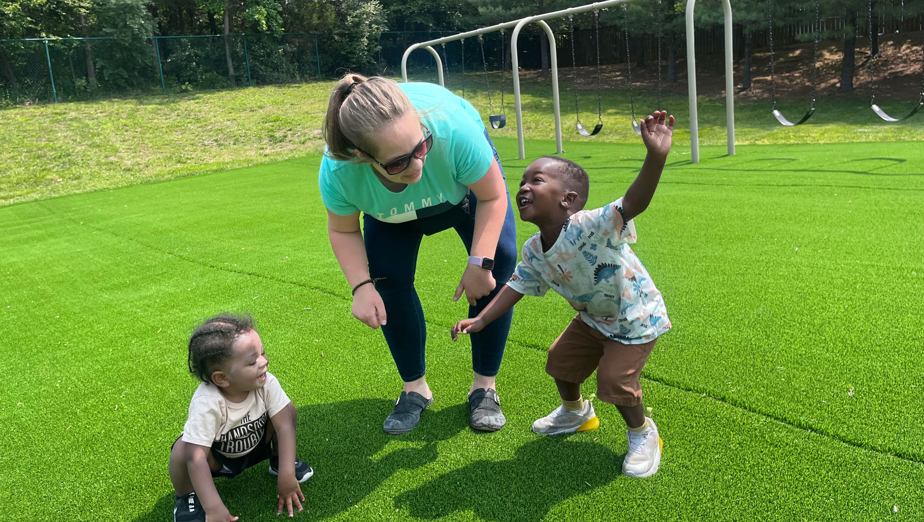 A teacher bending down to play with two laughing toddlers on the playground turf, one reaching up to her